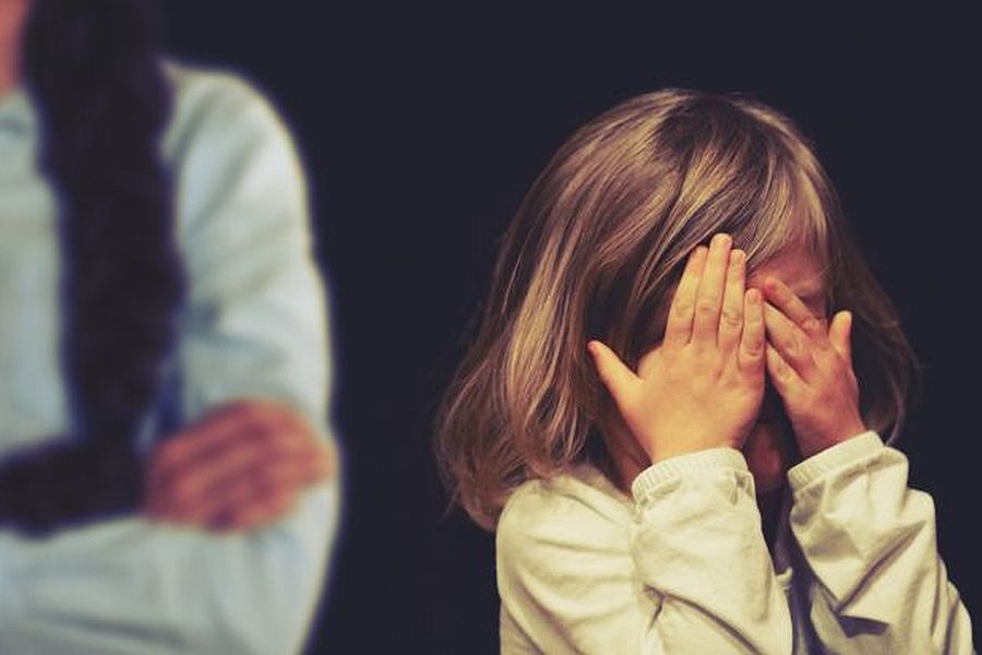 Ein kleines Mädchen hält sich die Augen zu, während im Hintergrund eine Frau mit verschränkten Armen zu sehen ist.